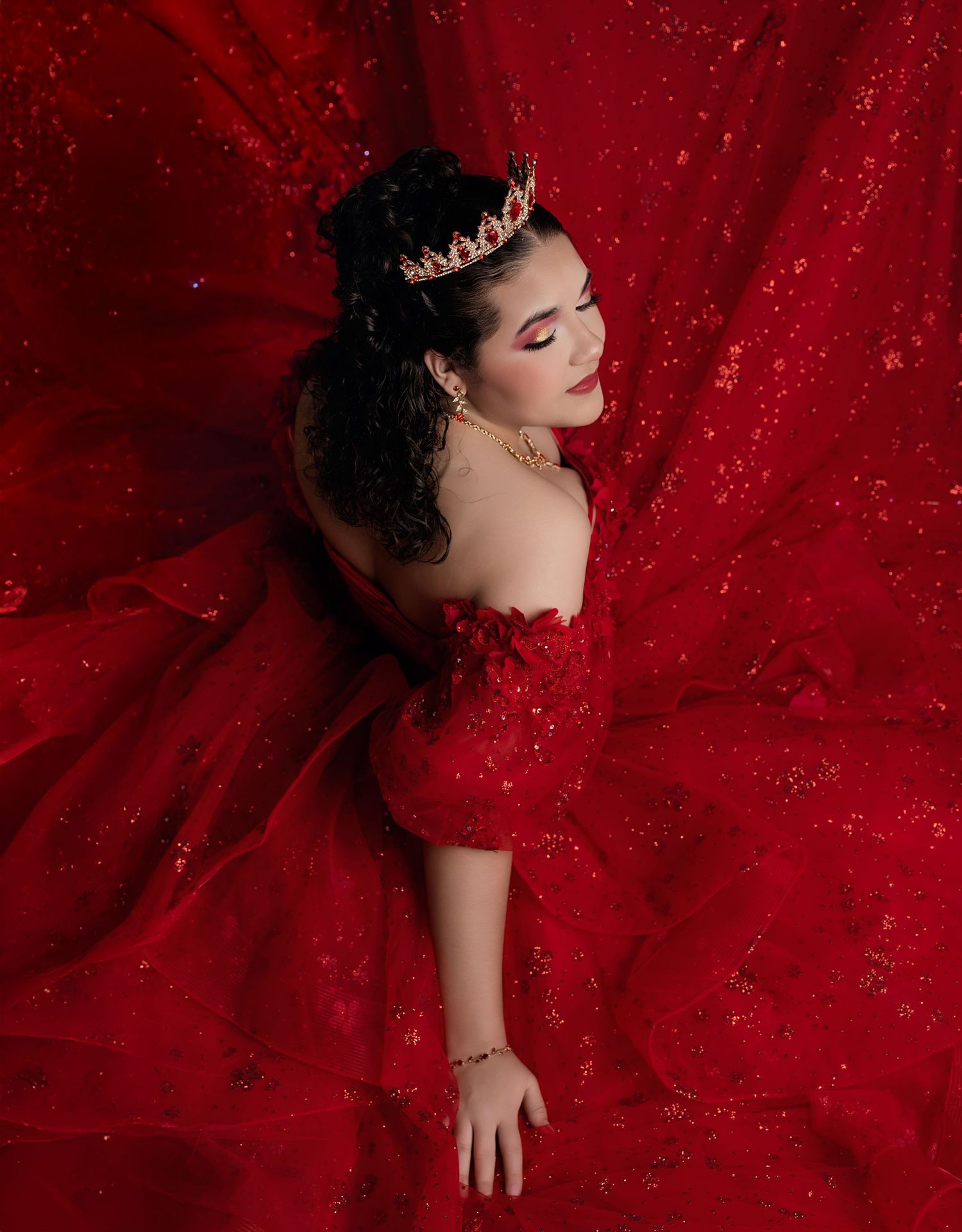 Young woman in red off-shoulder gown with crown poses elegantly against sparkling red fabric backdrop.