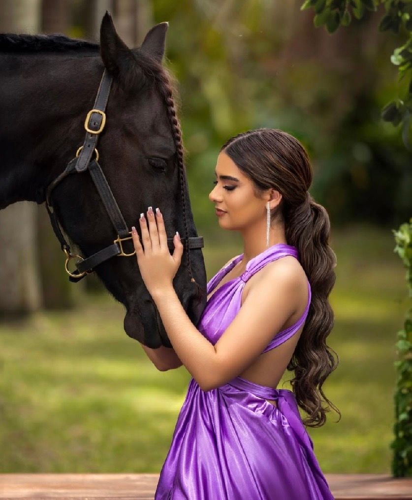 A young woman in a purple satin dress gently touches a black horse outdoors in a lush green setting.