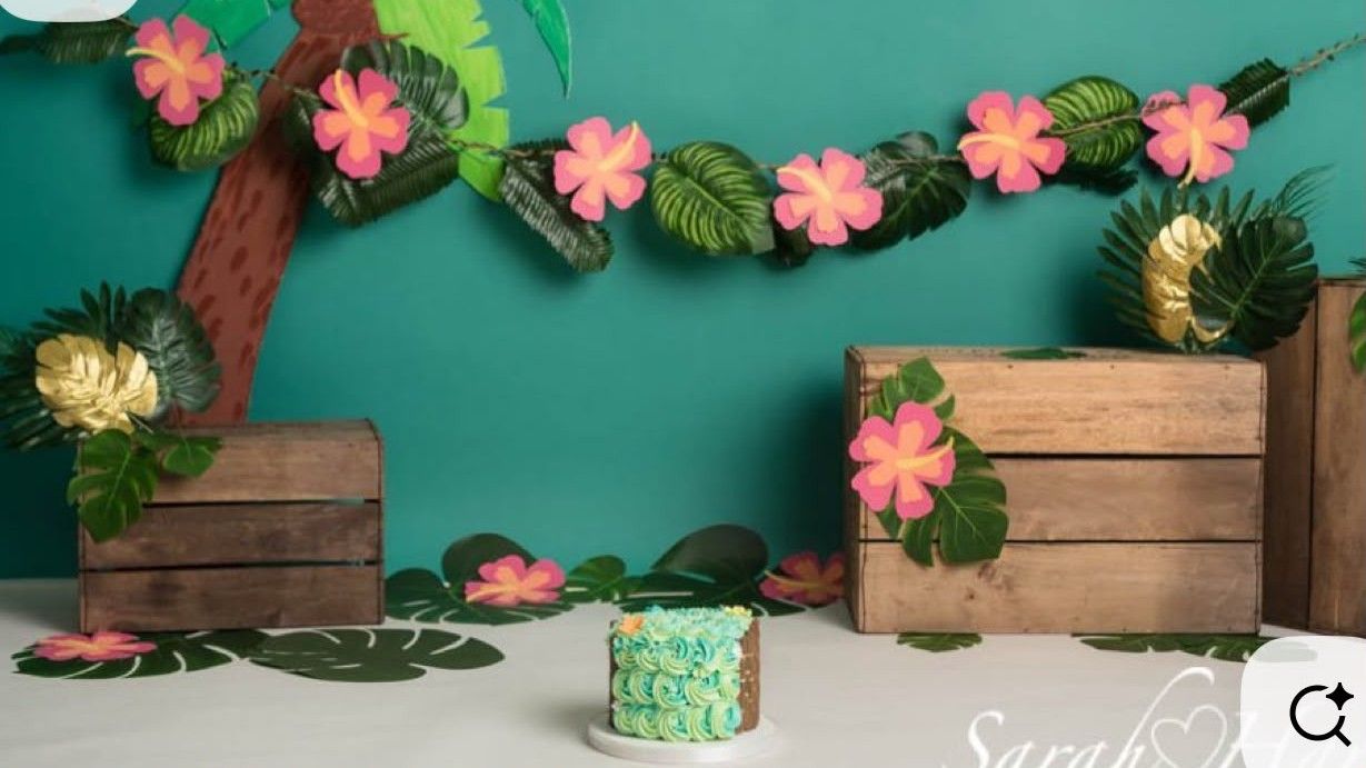Tropical themed cake smash setup with pink hibiscus garland, wooden crates, and a small green rosette cake on white floor.