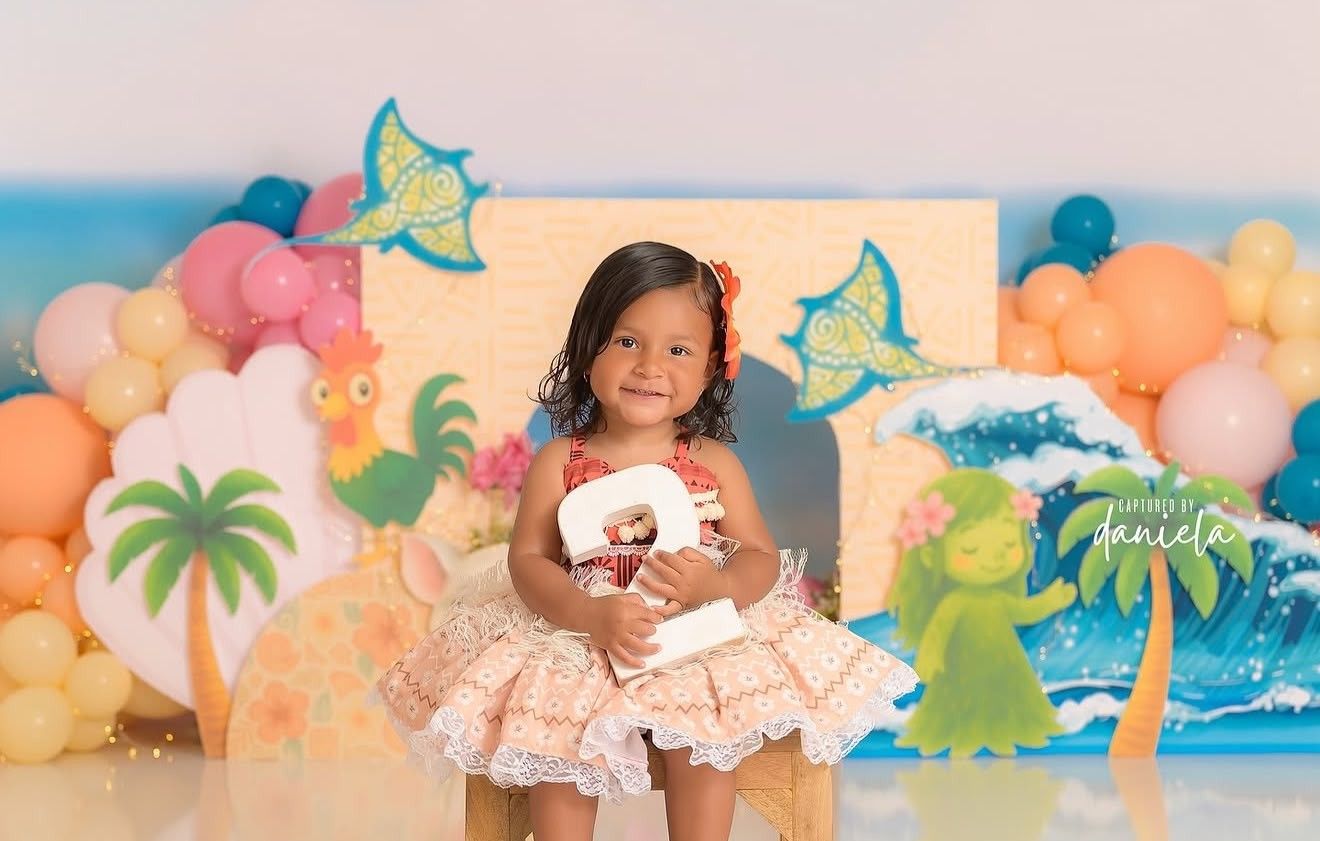 Toddler girl in a floral dress holds a doll at a tropical Moana-themed birthday party with balloon decorations.