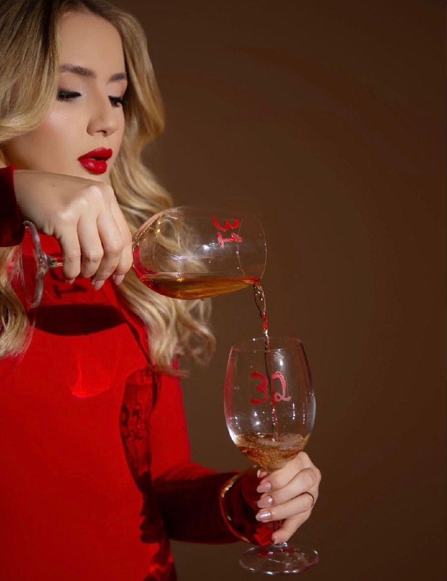 Blonde woman in red outfit pouring wine into a glass against a dark background.