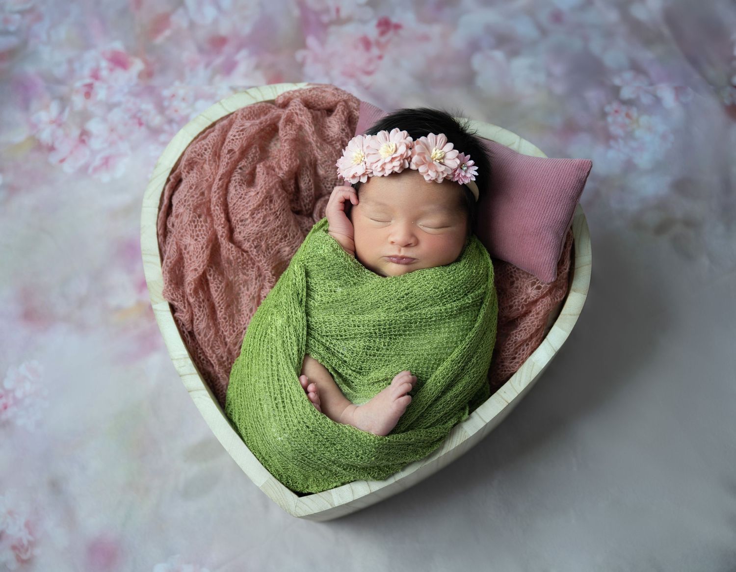 Newborn sleeping in heart-shaped basket with green wrap and pink accents.