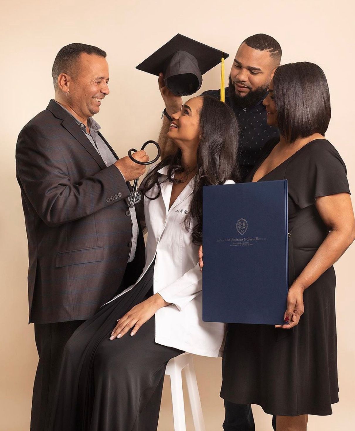 A graduate in white holds a diploma while celebrating with loved ones in formal attire during a graduation photoshoot.
