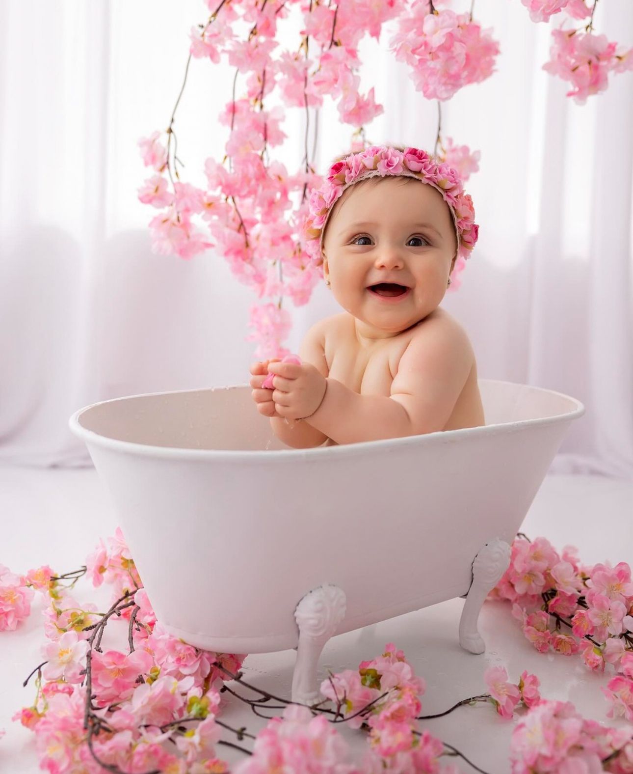 A baby wearing a pink floral bonnet sits in a white bathtub surrounded by pink cherry blossom branches.