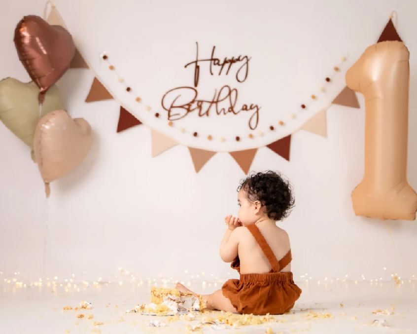 First birthday cake smash photo setup with rose gold balloons, bunting, and scattered cake crumbs on cream backdrop.