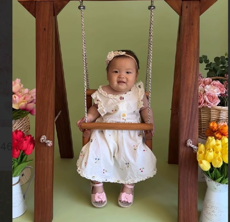 A baby wearing a white dress sits on a wooden porch swing, surrounded by colorful spring flowers and decorations.