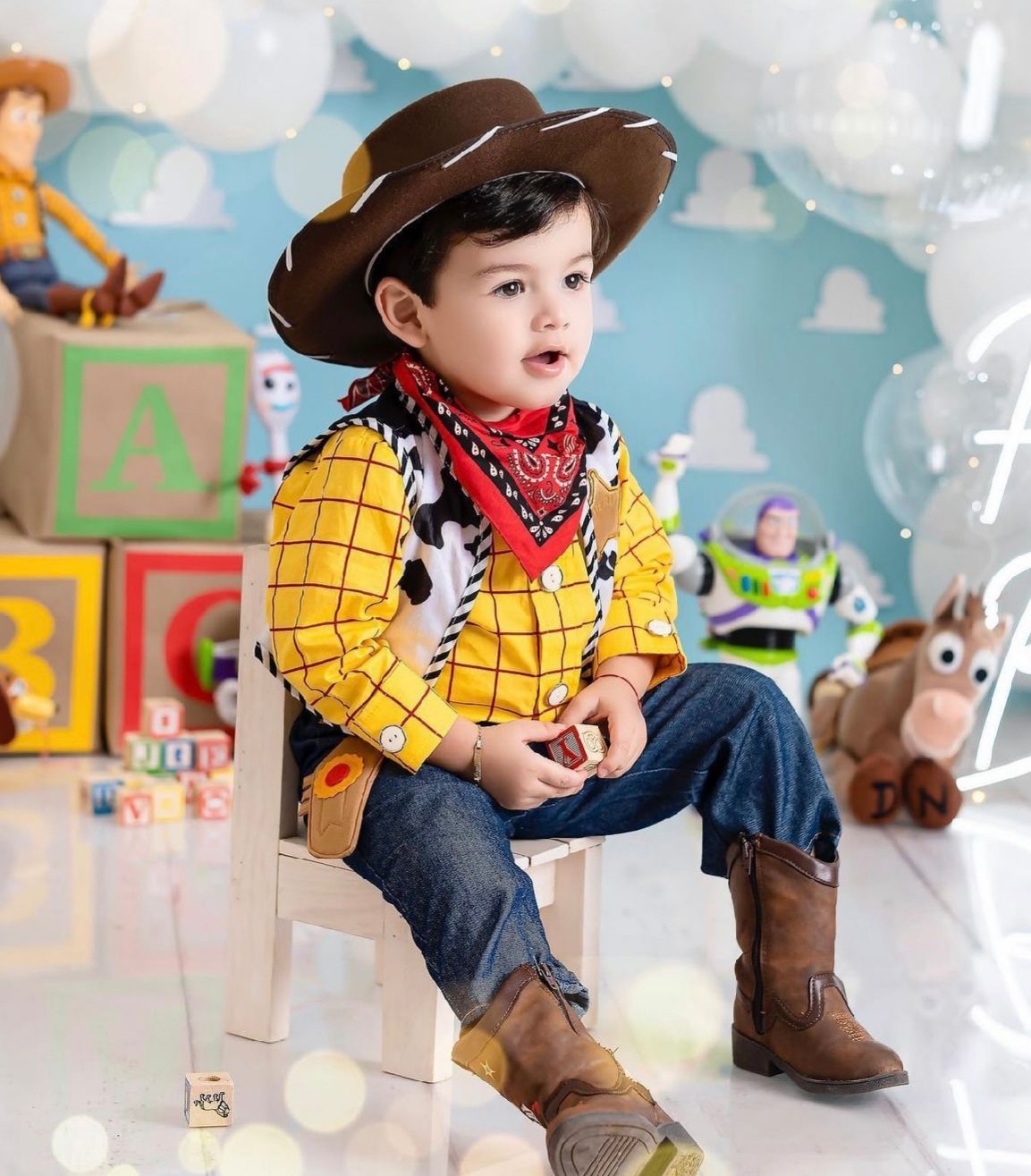 A small child wearing a yellow cowboy costume and brown boots sits on a white floor in a playful setting.