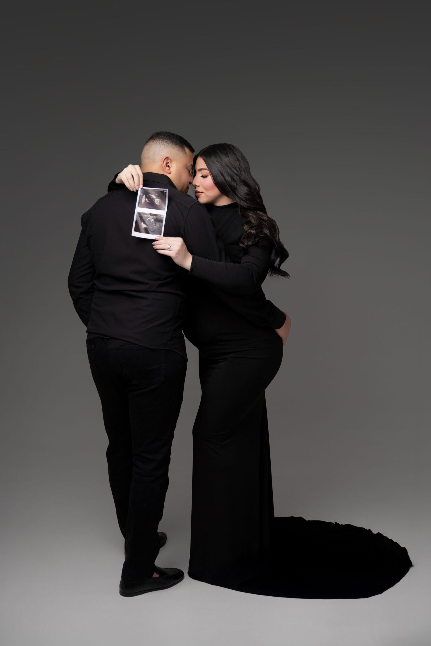 Couple in black formal wear embracing, woman in elegant gown holding ultrasound photo, maternity portrait on gray background.