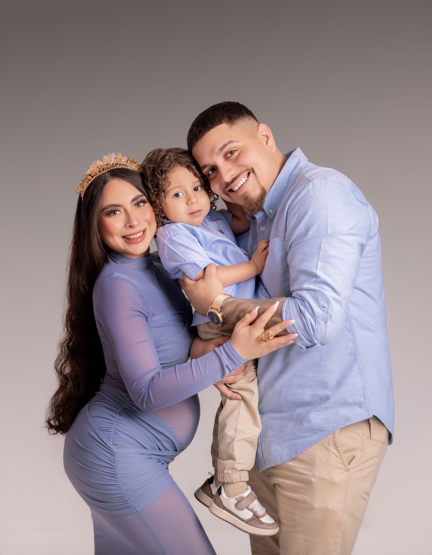 Pregnant woman in blue dress poses with smiling man and toddler son in coordinating blue outfits for maternity photo.