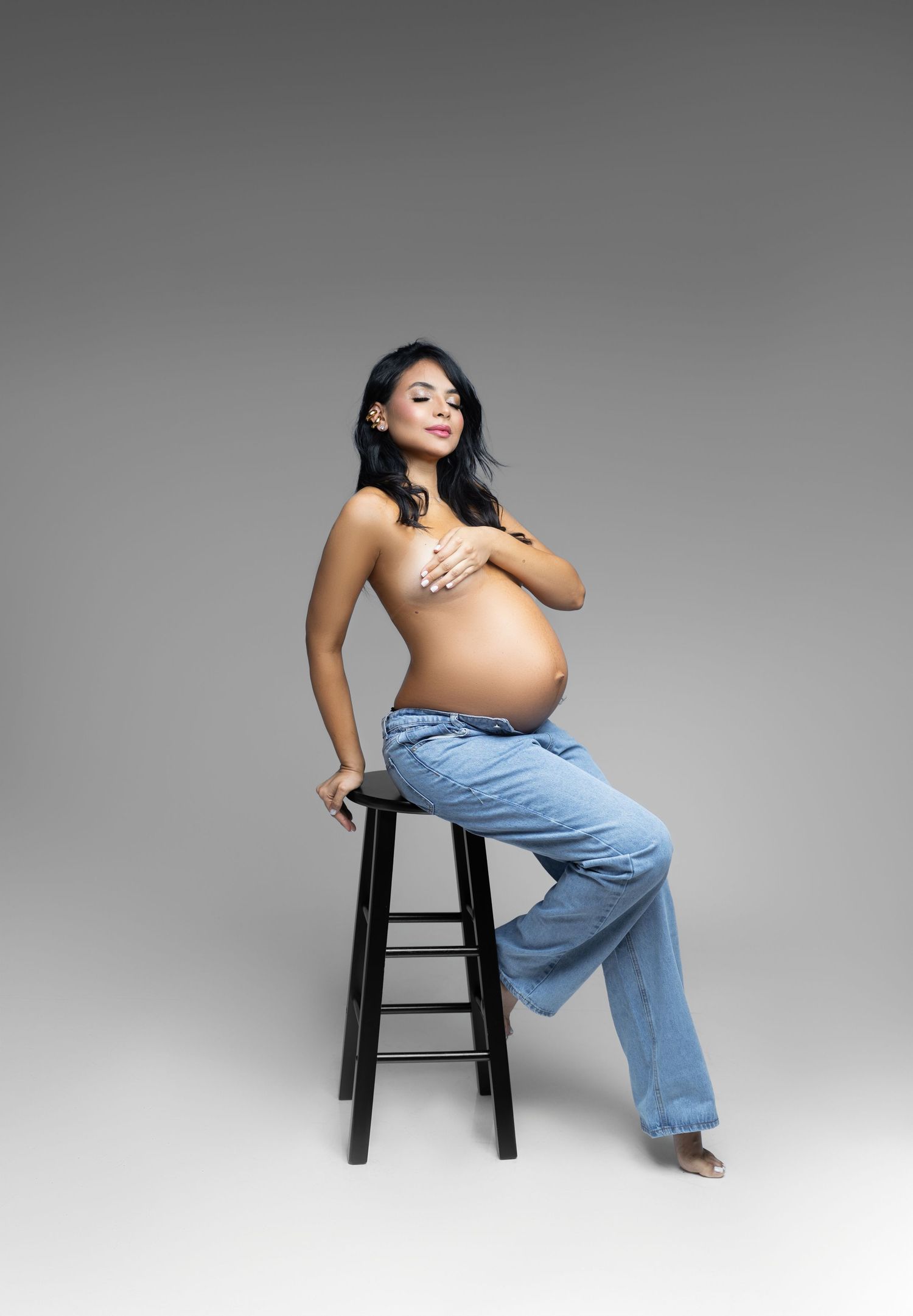 Pregnant woman sitting on a stool, wearing blue jeans, posing for a maternity photo shoot in a studio.