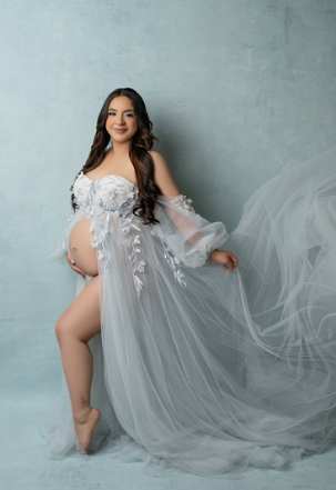 Artistic maternity photoshoot featuring ethereal white sheer gown with floral details against a grey textured backdrop.