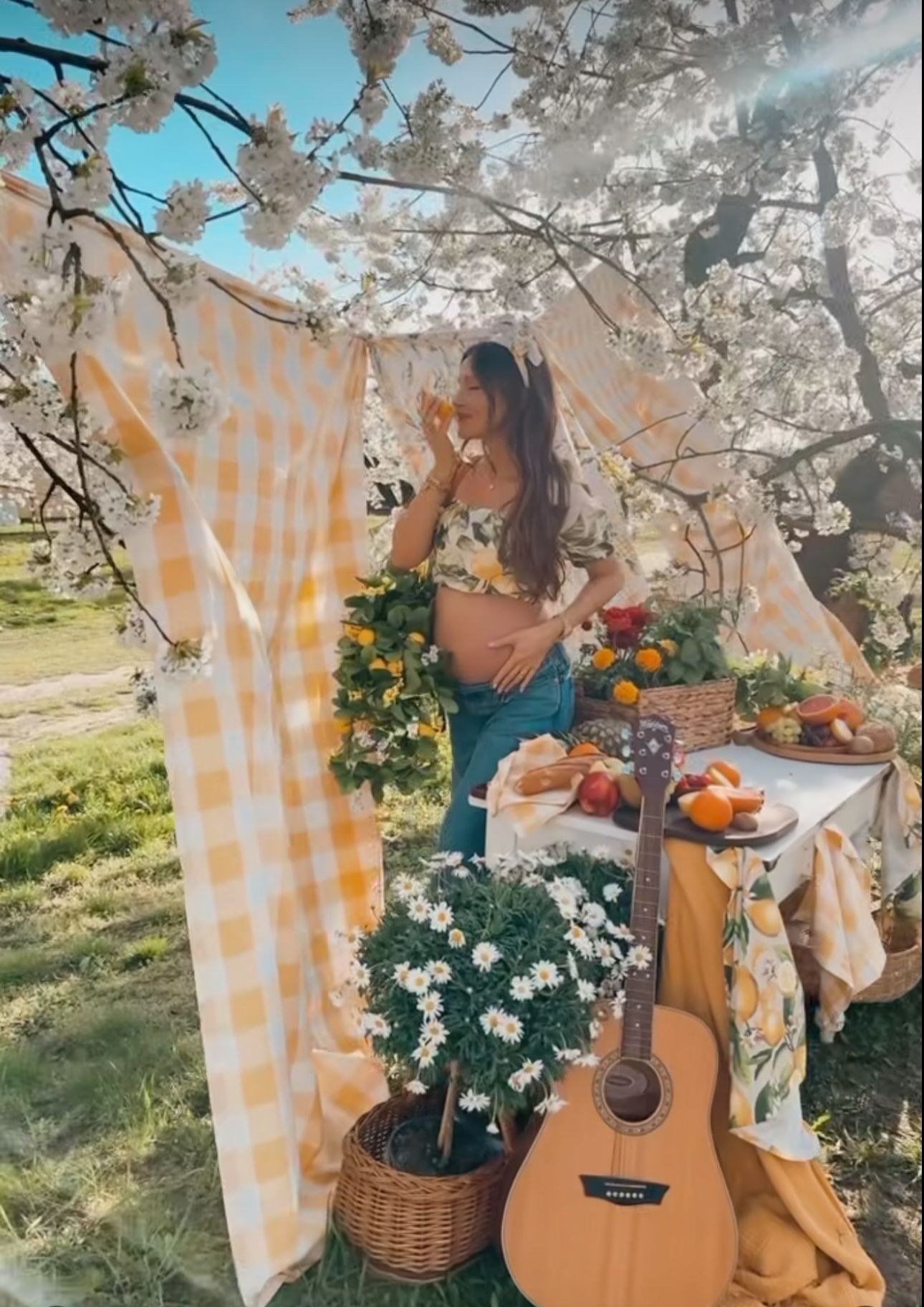 Pregnant woman in boho outdoor setup with guitar, flowers, fruits, yellow gingham curtains, and cherry blossoms.