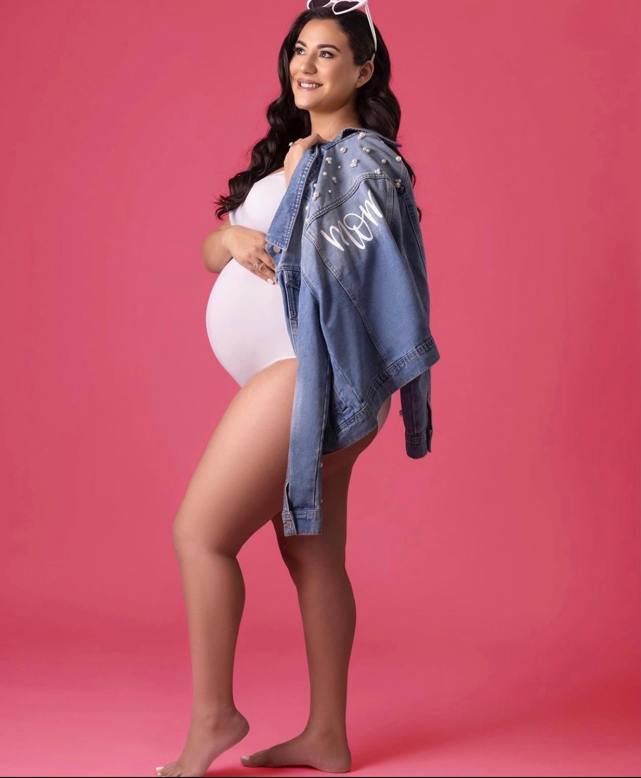Smiling pregnant woman in white bodysuit and denim jacket posing against a pink background.
