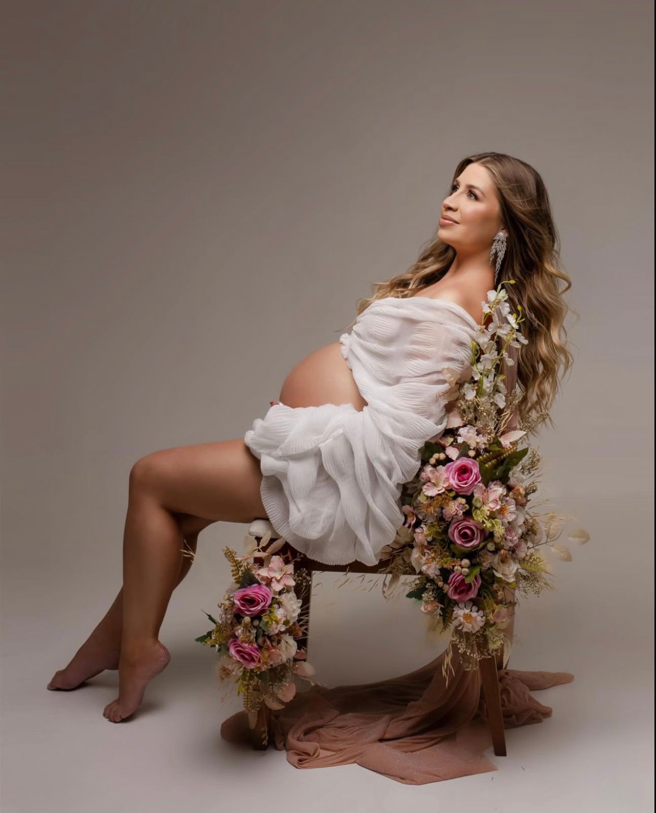 Pregnant woman in white draped fabric sits on floral-adorned chair in elegant studio maternity portrait.