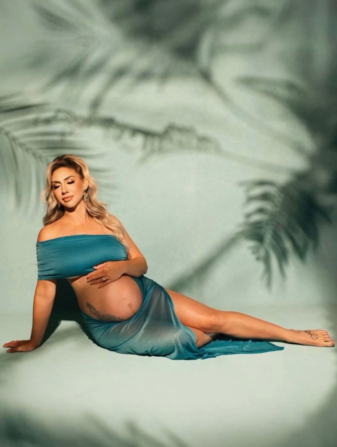 Pregnant woman in teal off-shoulder gown poses gracefully on floor with palm leaf shadows in maternity photoshoot.