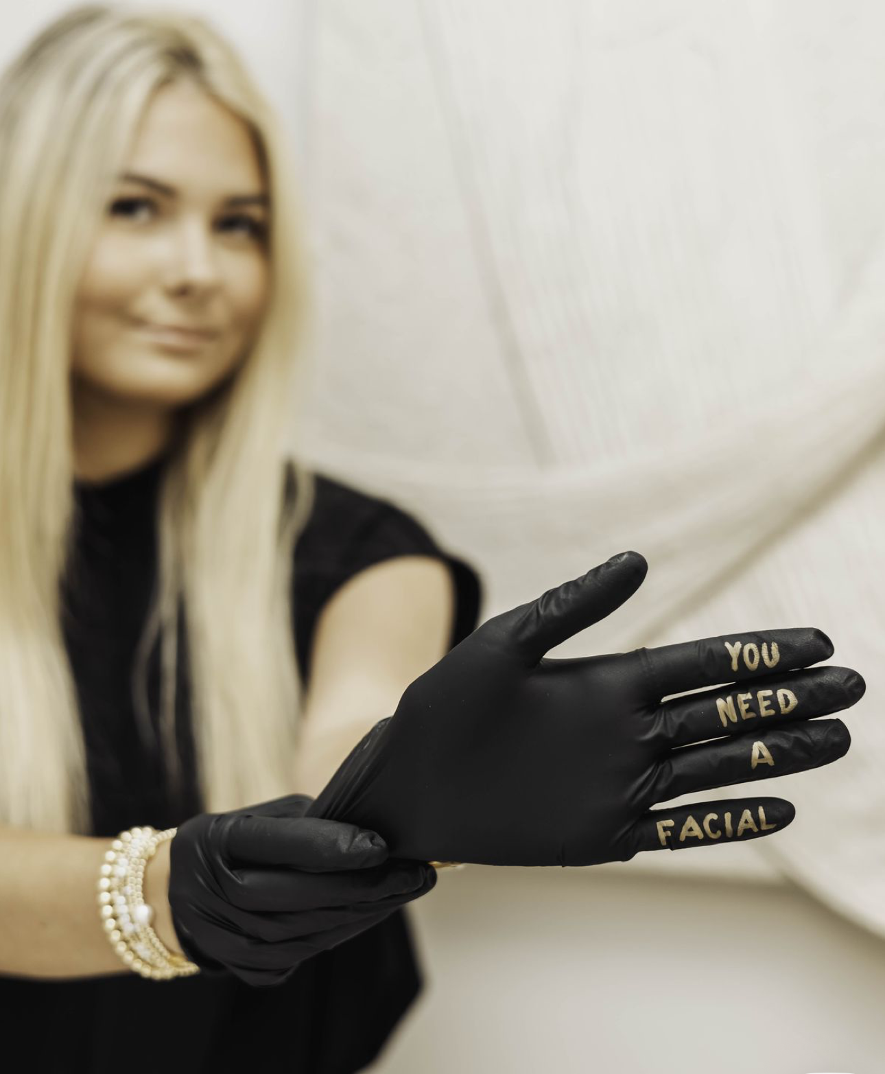 Woman in black gloves with gold text reading YOU NEED A FACIAL, promoting skincare services.