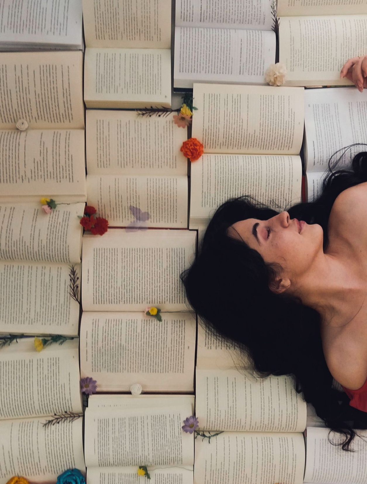 Woman lying among open books with colorful flowers, viewed from above in an artistic literary-themed portrait.
