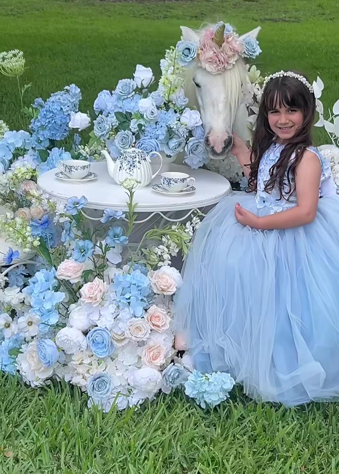 Young girl in blue tulle dress poses with a white horse and floral tea party setup outdoors.