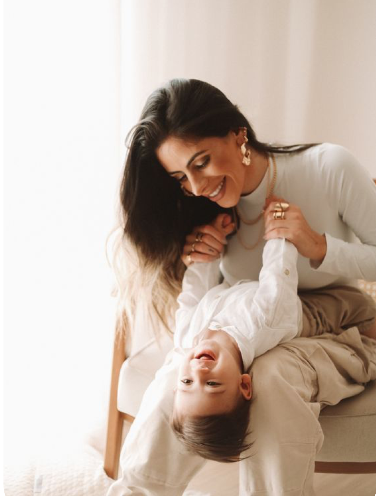 A smiling woman with dark hair holds a laughing child upside down in a bright, airy room, both wearing white.