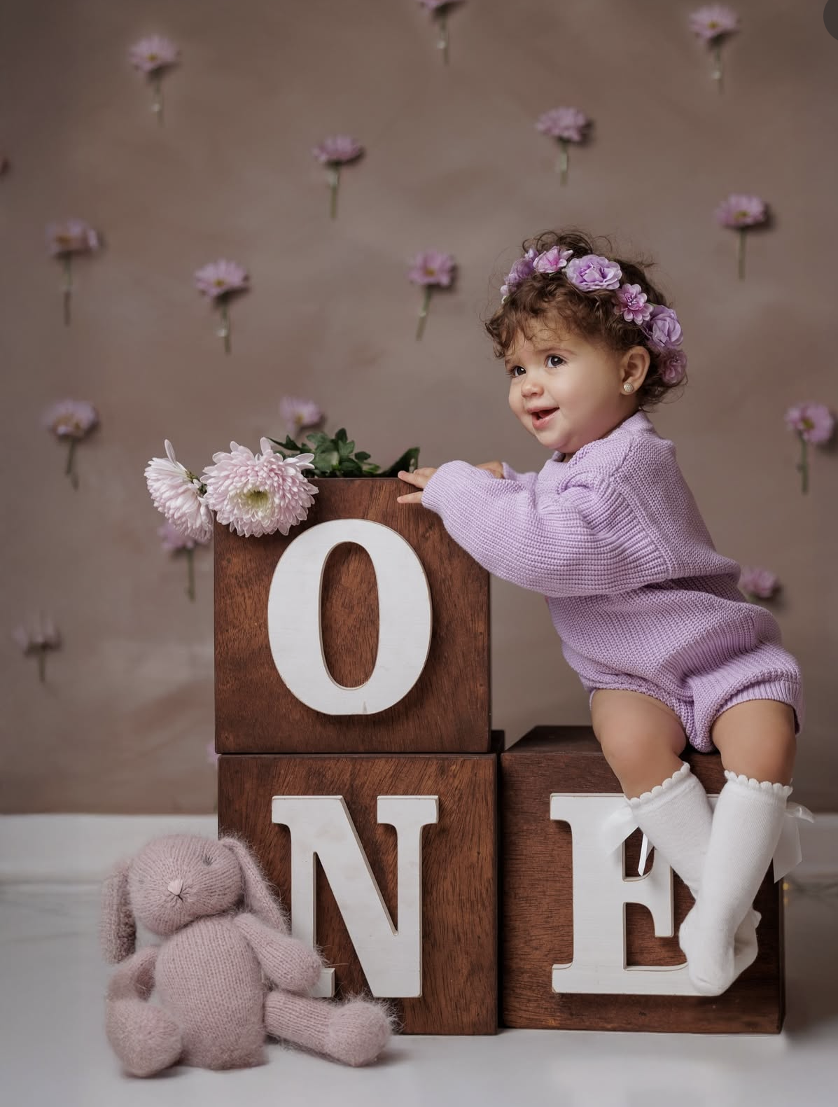 Baby girl in purple romper and floral crown poses with wooden ONE blocks and pink bunny for first birthday photo.