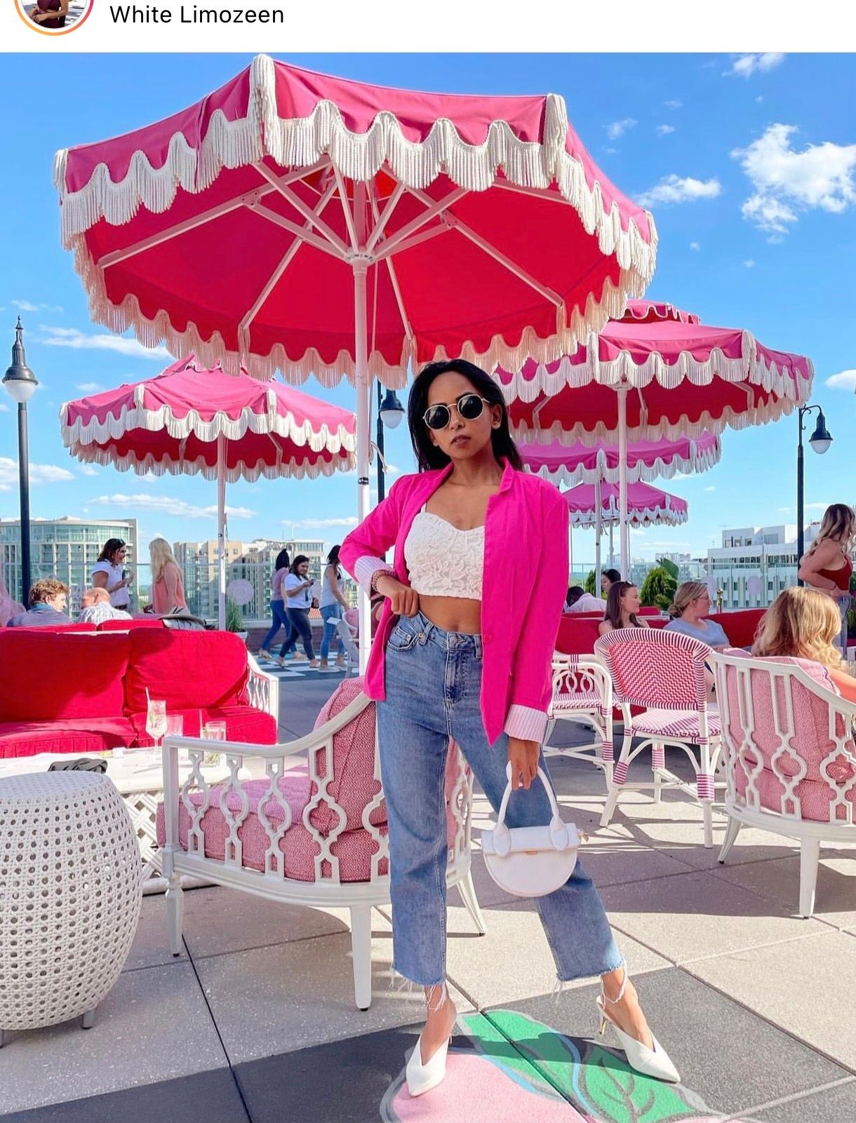 Woman in pink blazer and jeans poses on rooftop patio with vibrant pink fringed umbrellas at White Limozeen.