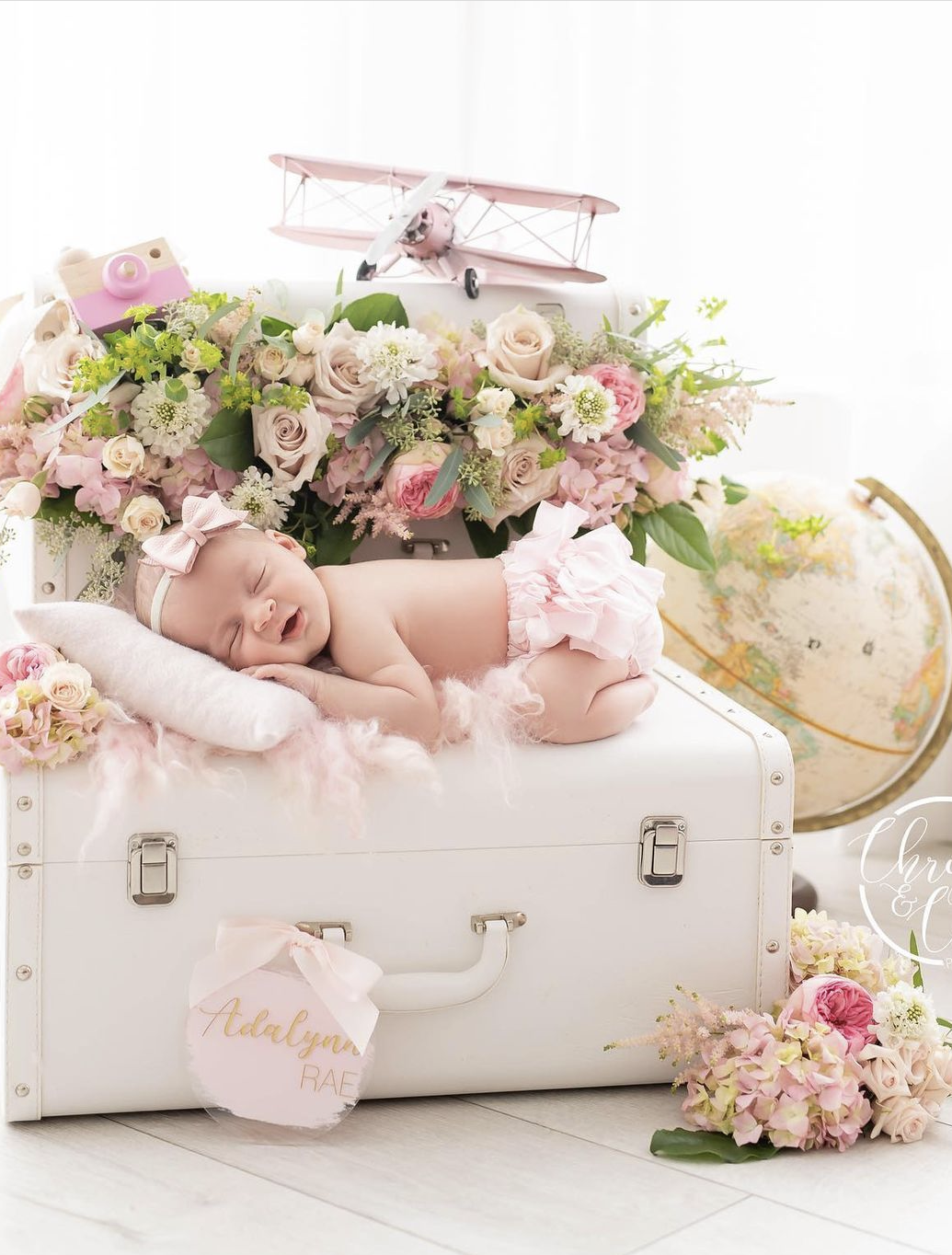 Sleeping newborn baby in pink bow outfit on white vintage suitcase with floral arrangement and globe.