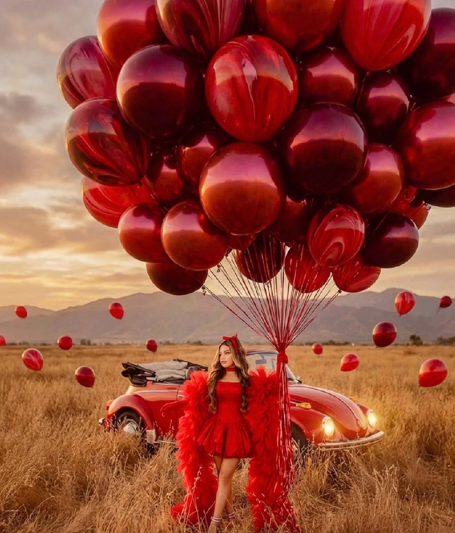 Woman in red dress holds large bundle of red balloons in golden field with vintage red car at sunset.