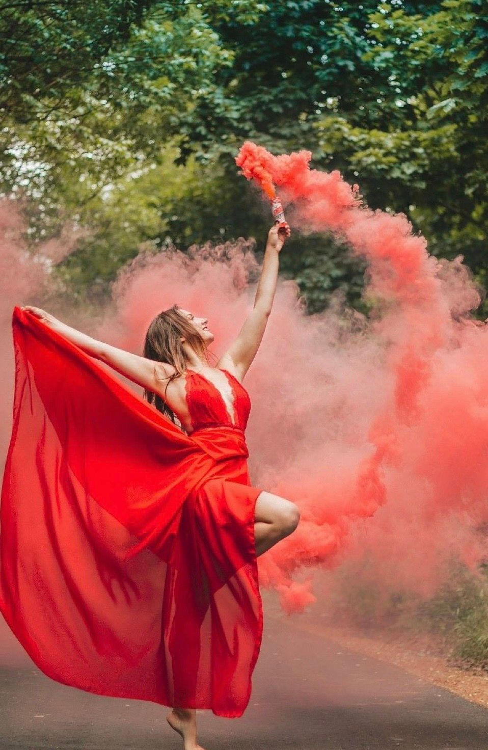 Woman in flowing red dress dancing on road holding red smoke flare surrounded by colorful smoke in lush green setting.