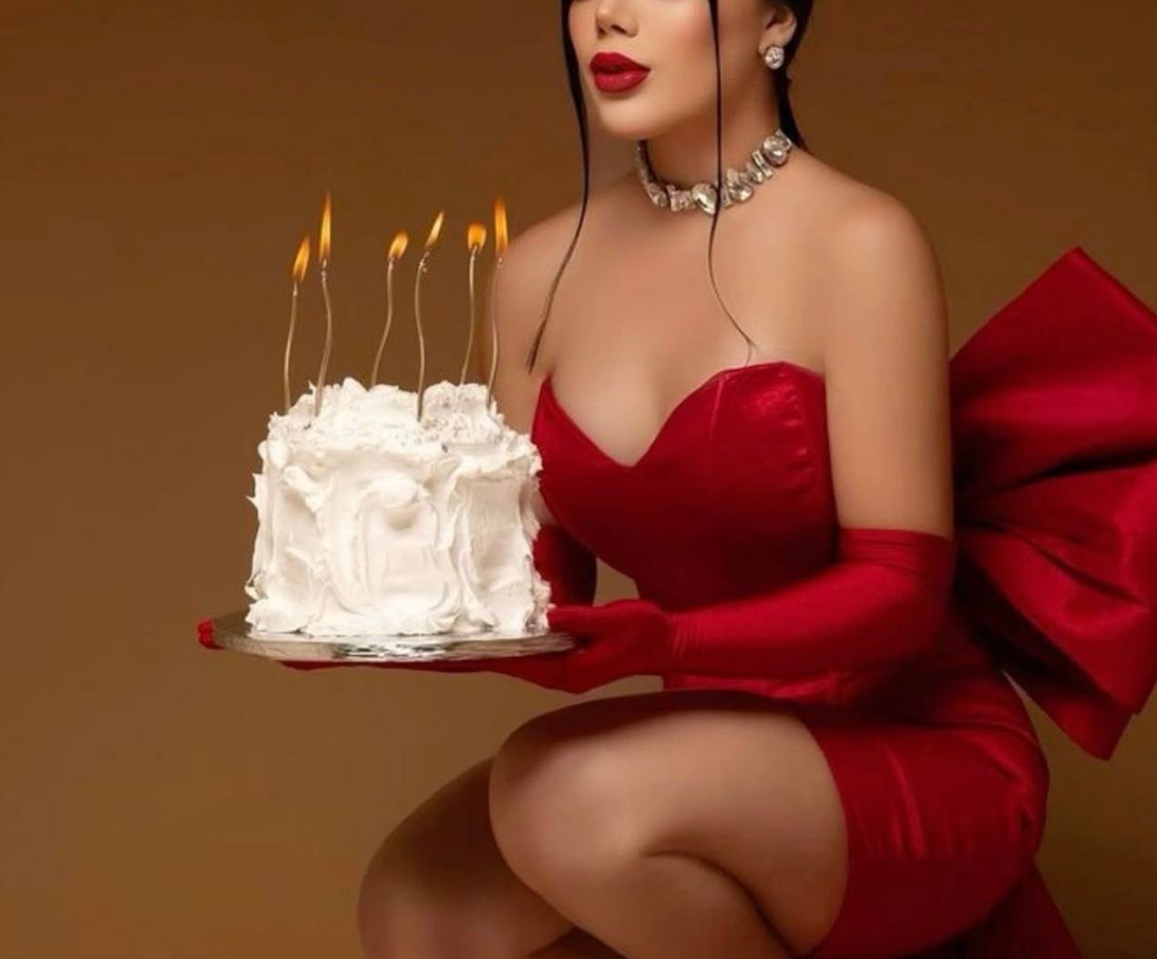Woman in red strapless dress and gloves holding white birthday cake with lit candles against brown background.