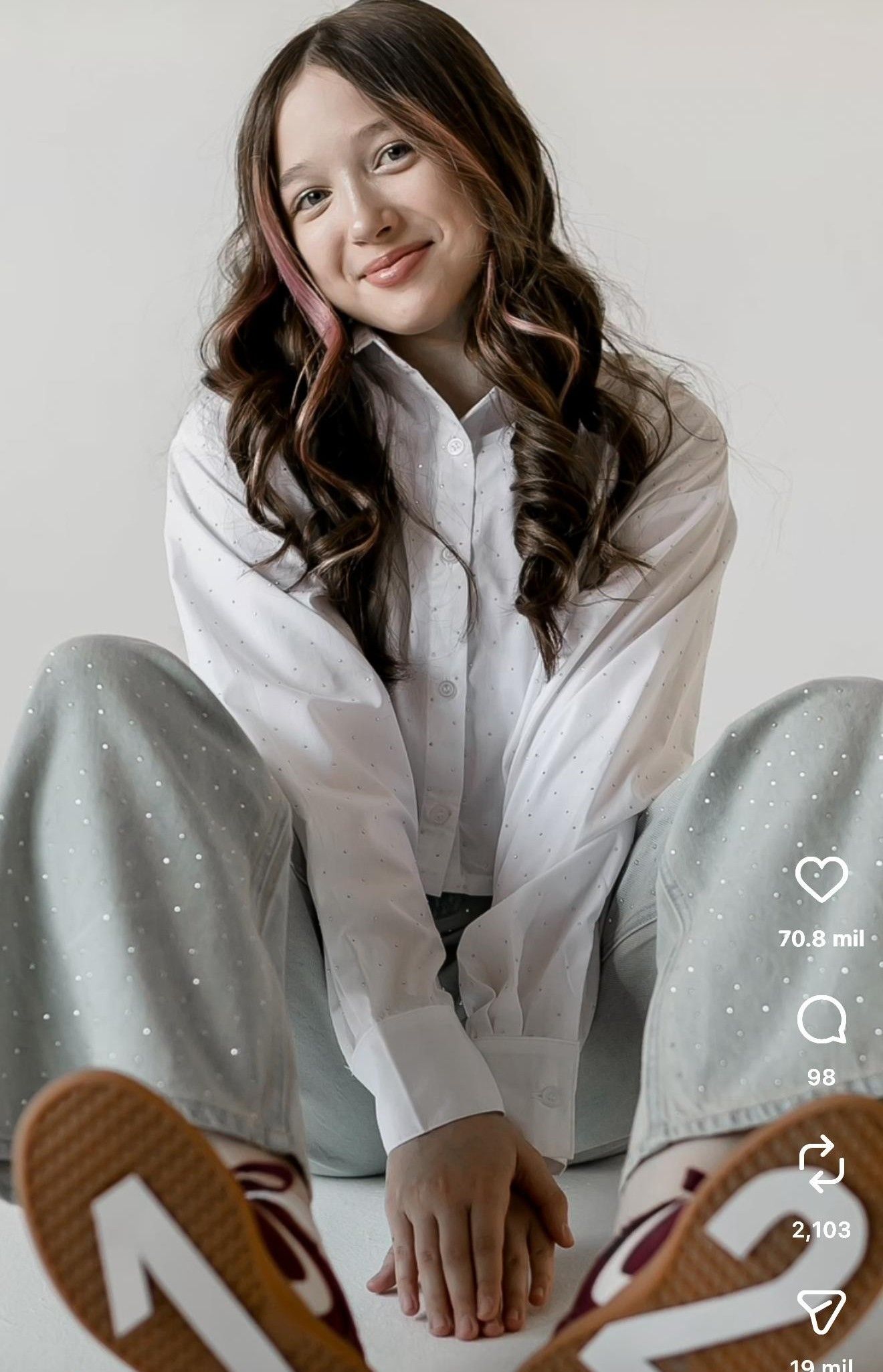 Young woman with curly brown hair smiling, wearing a white shirt and light blue sparkle pants, sitting casually.