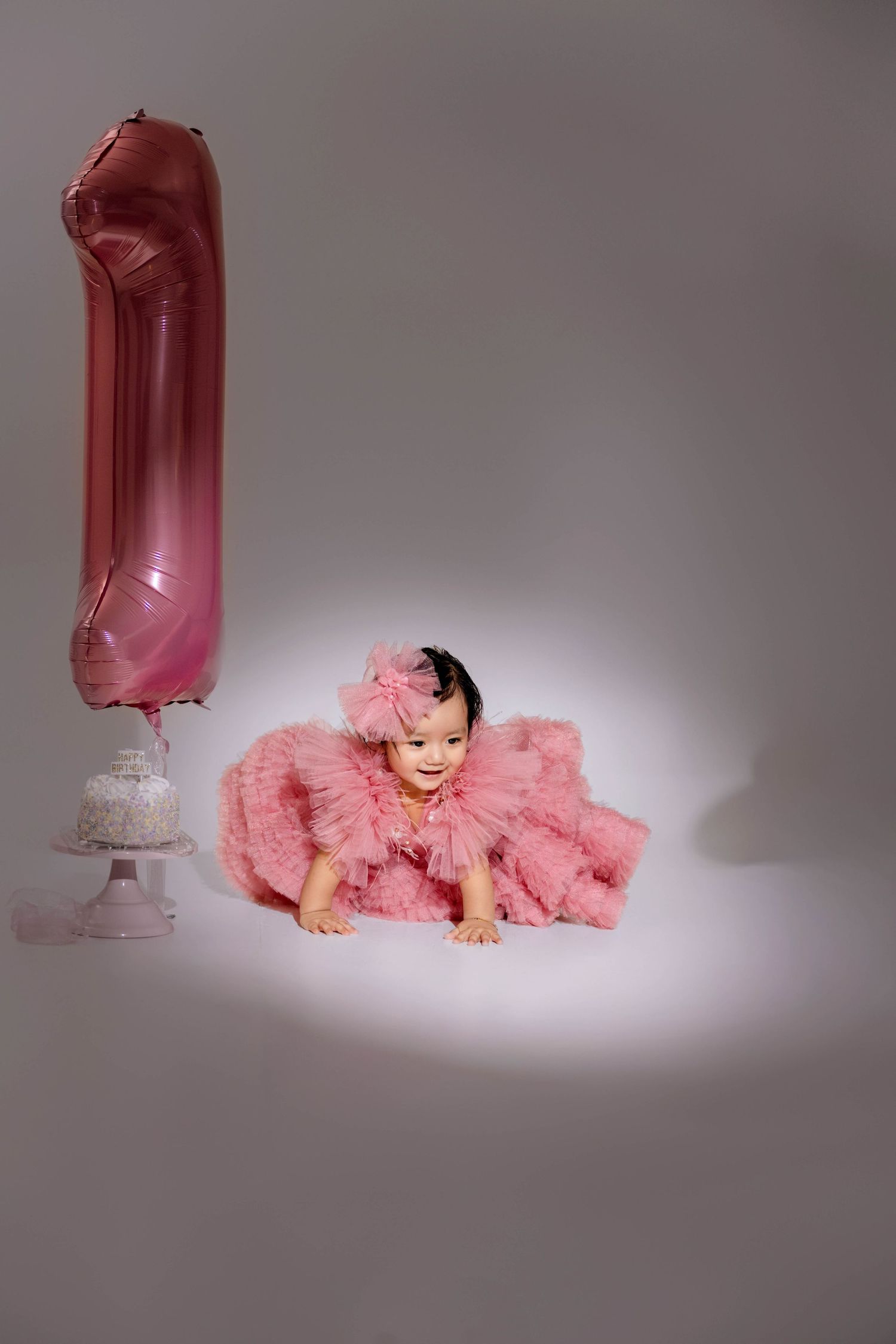 Baby girl in pink tutu dress crawls near number 1 balloon and cake celebrating her first birthday.