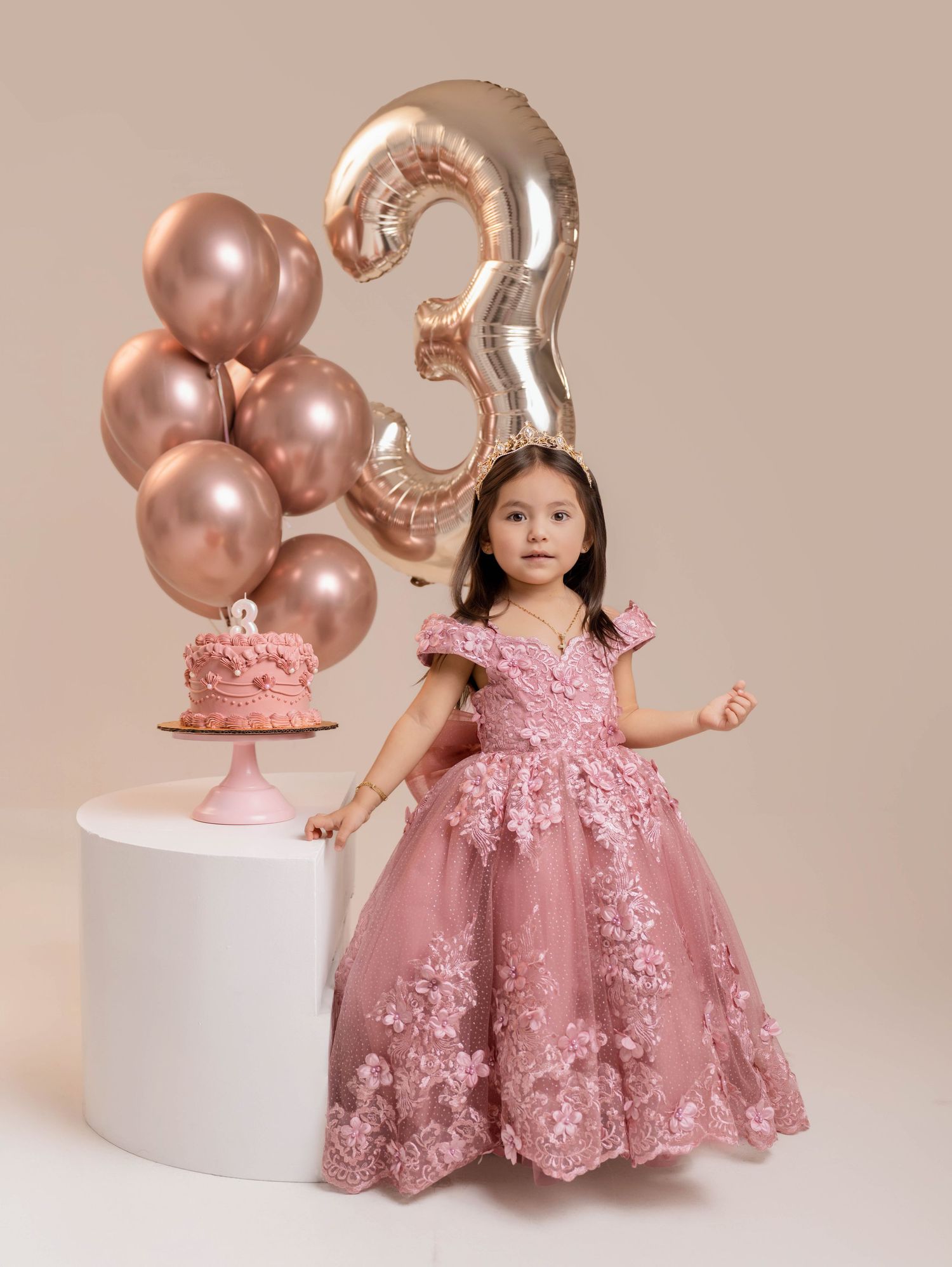 A young girl in a pink floral ball gown poses beside a birthday cake and balloons celebrating her 3rd birthday.