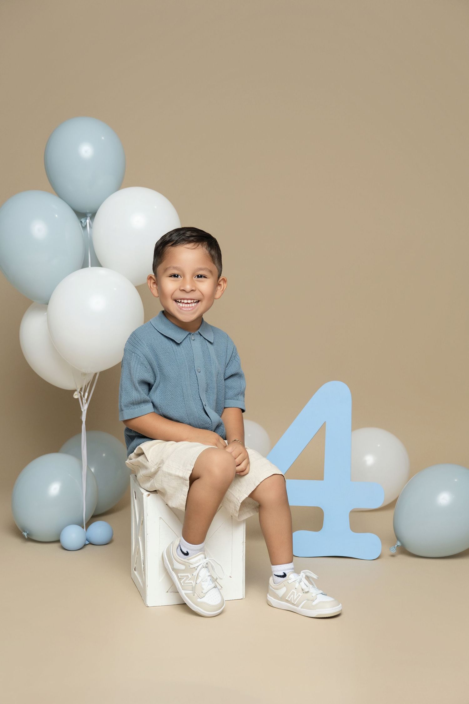 A smiling boy sits on a white box beside a blue number 4 and blue and white balloons, celebrating his 4th birthday.
