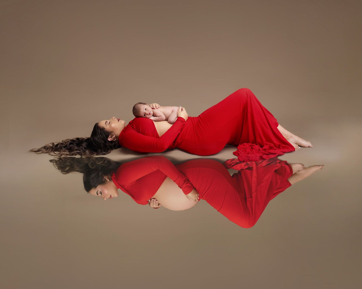 Woman in red gown lying on reflective floor holding newborn baby in a elegant maternity studio portrait.
