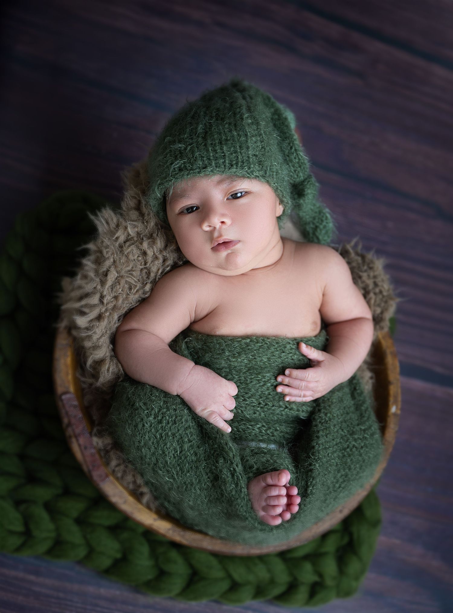 Newborn baby wearing a green knit hat and wrap, resting peacefully in a wooden bowl on a dark wood surface.