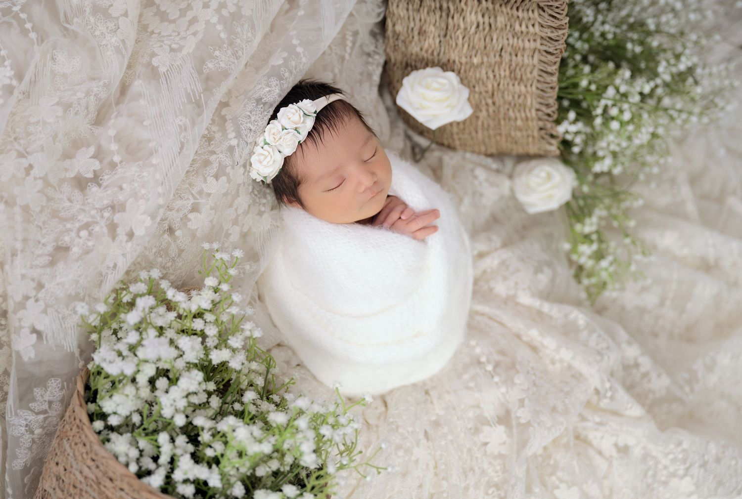 Sleeping newborn baby wrapped in white, wearing a lace headband, surrounded by white flowers and burlap baskets.