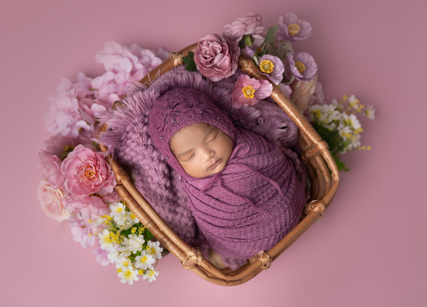 Newborn baby swaddled in purple wrap sleeping in a wicker basket surrounded by pink and white flowers.