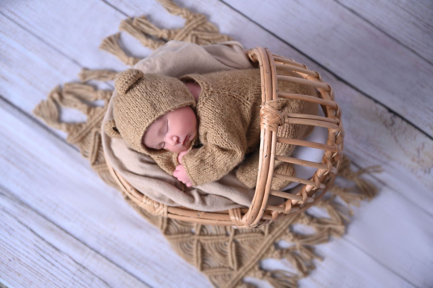 Newborn baby sleeping peacefully in a wicker basket with beige knit wrap and hat on white wood floor.
