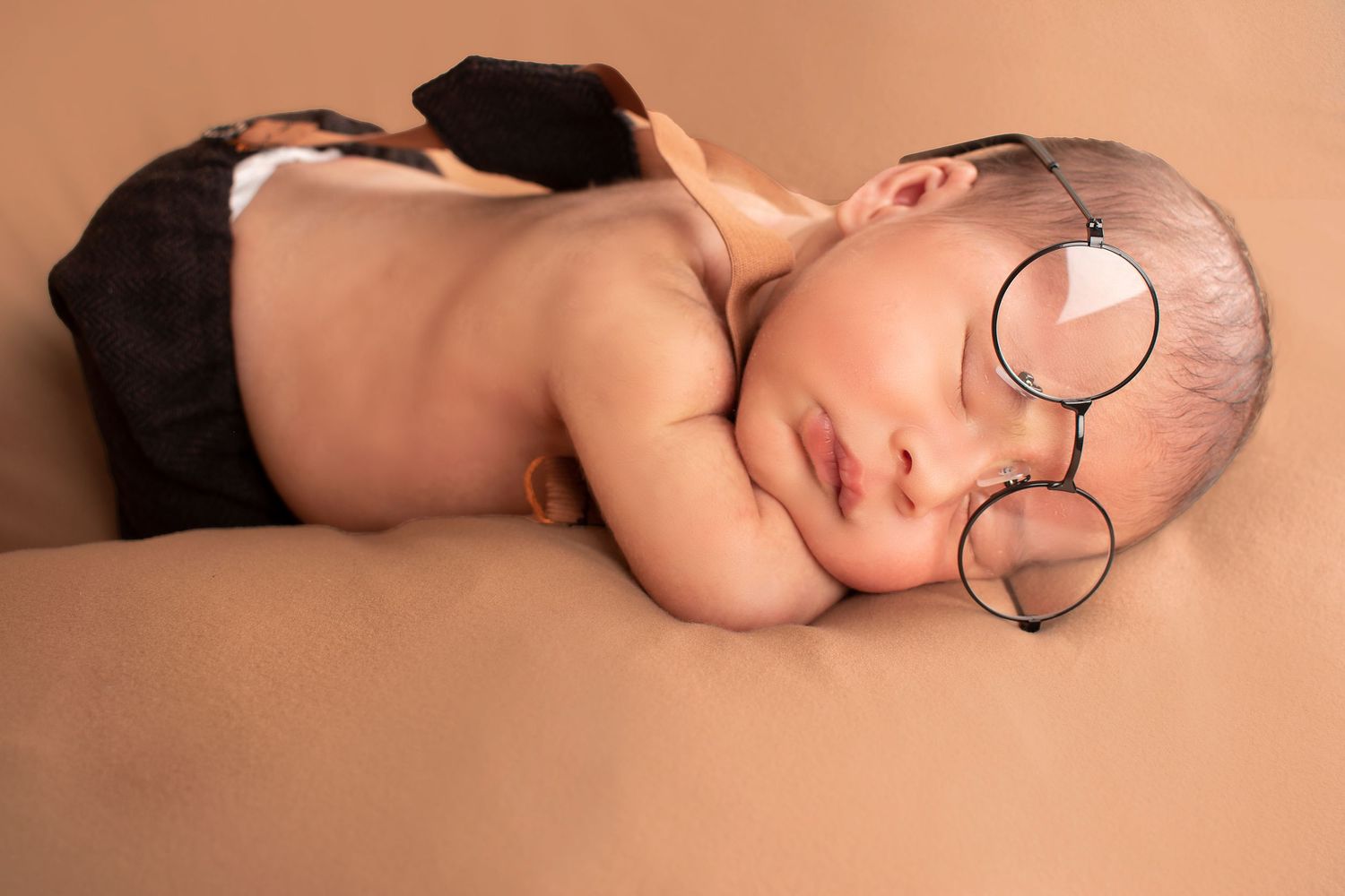 Adorable sleeping newborn baby wearing tiny round glasses and dark suspenders on a warm beige background.