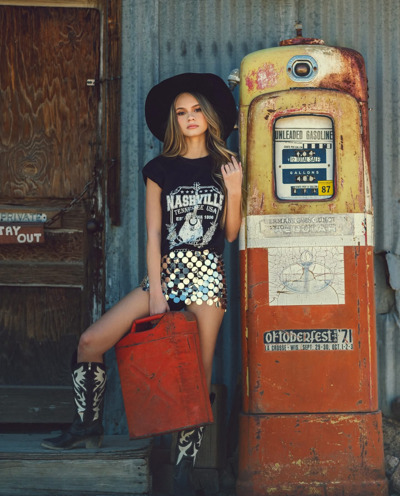 Young woman in Nashville tee, sequin shorts, cowboy boots and hat poses by vintage rusty gas pump holding red fuel can.