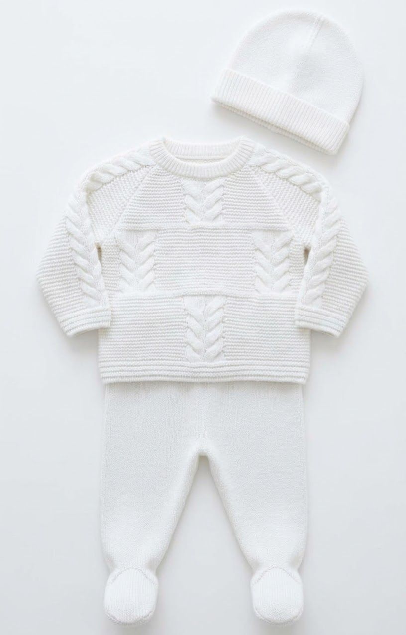 White knit baby 3-piece set featuring cable-knit sweater, footed pants with pom-poms, and matching beanie hat.