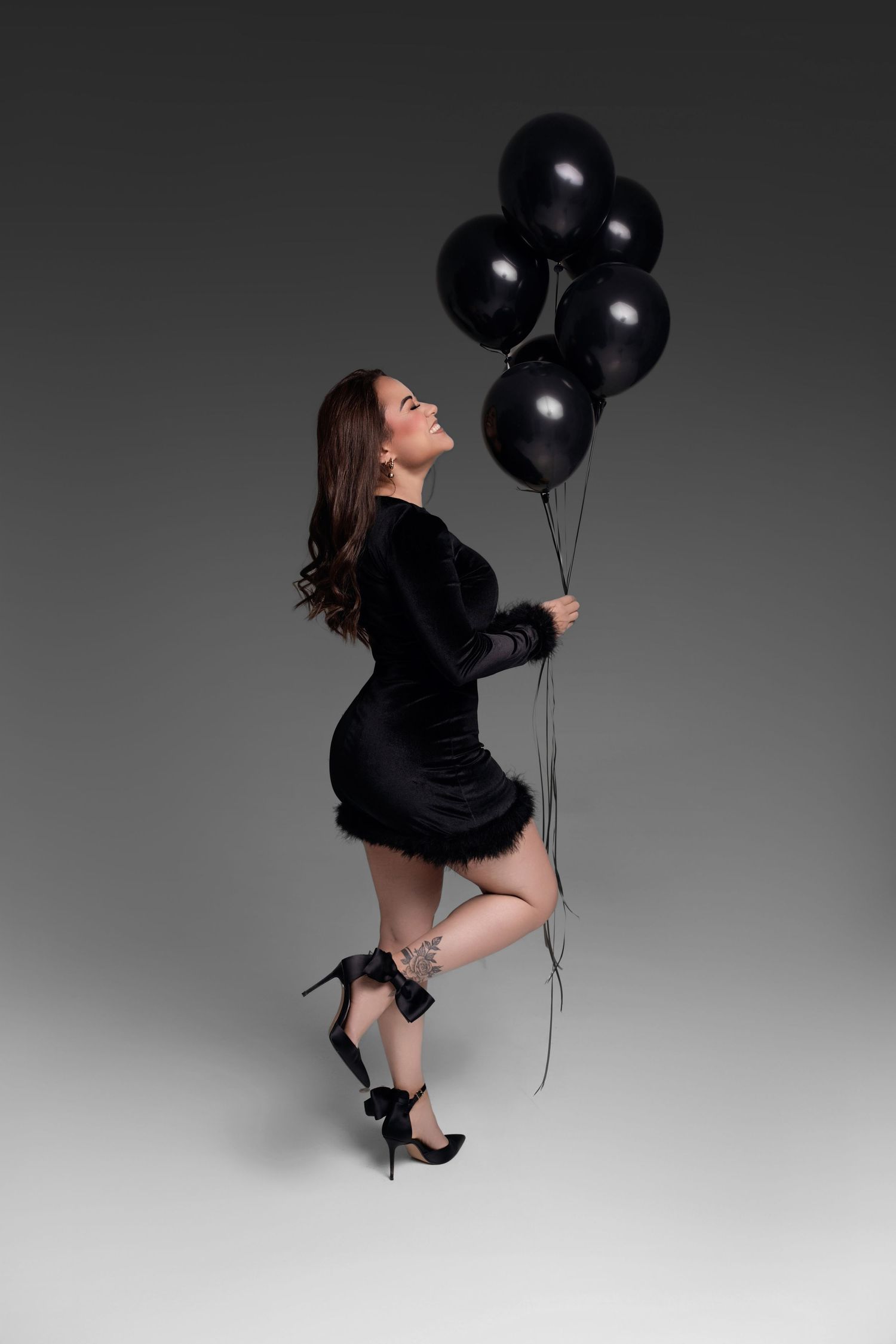 Woman in black velvet mini dress and heels holds black balloons, posing elegantly against gray background.