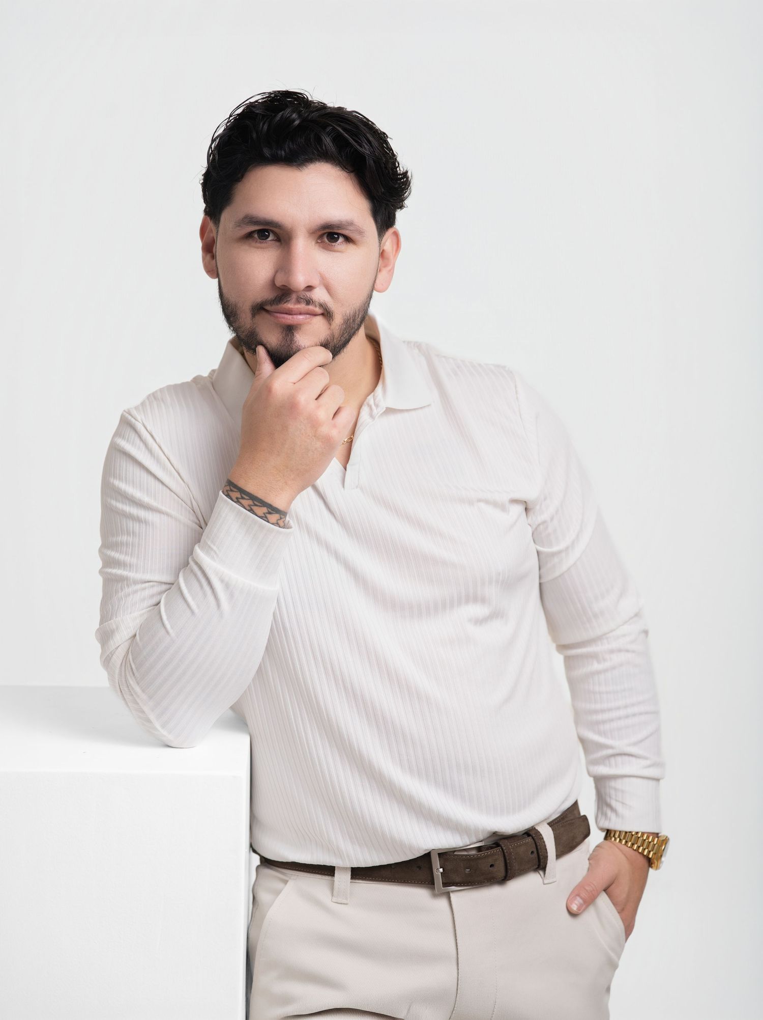 Man with dark styled hair and a beard poses thoughtfully with hand on chin, wearing a cream ribbed shirt, brown belt and gold watch.