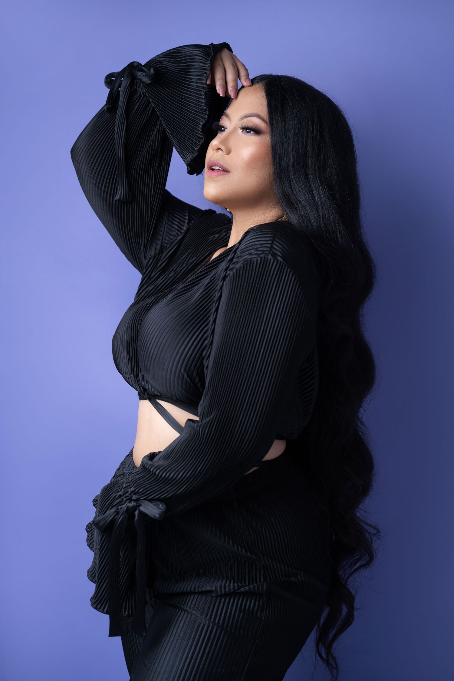 Woman with long wavy black hair poses with one arm raised in a black pleated bell-sleeve top and pants against a purple studio backdrop.