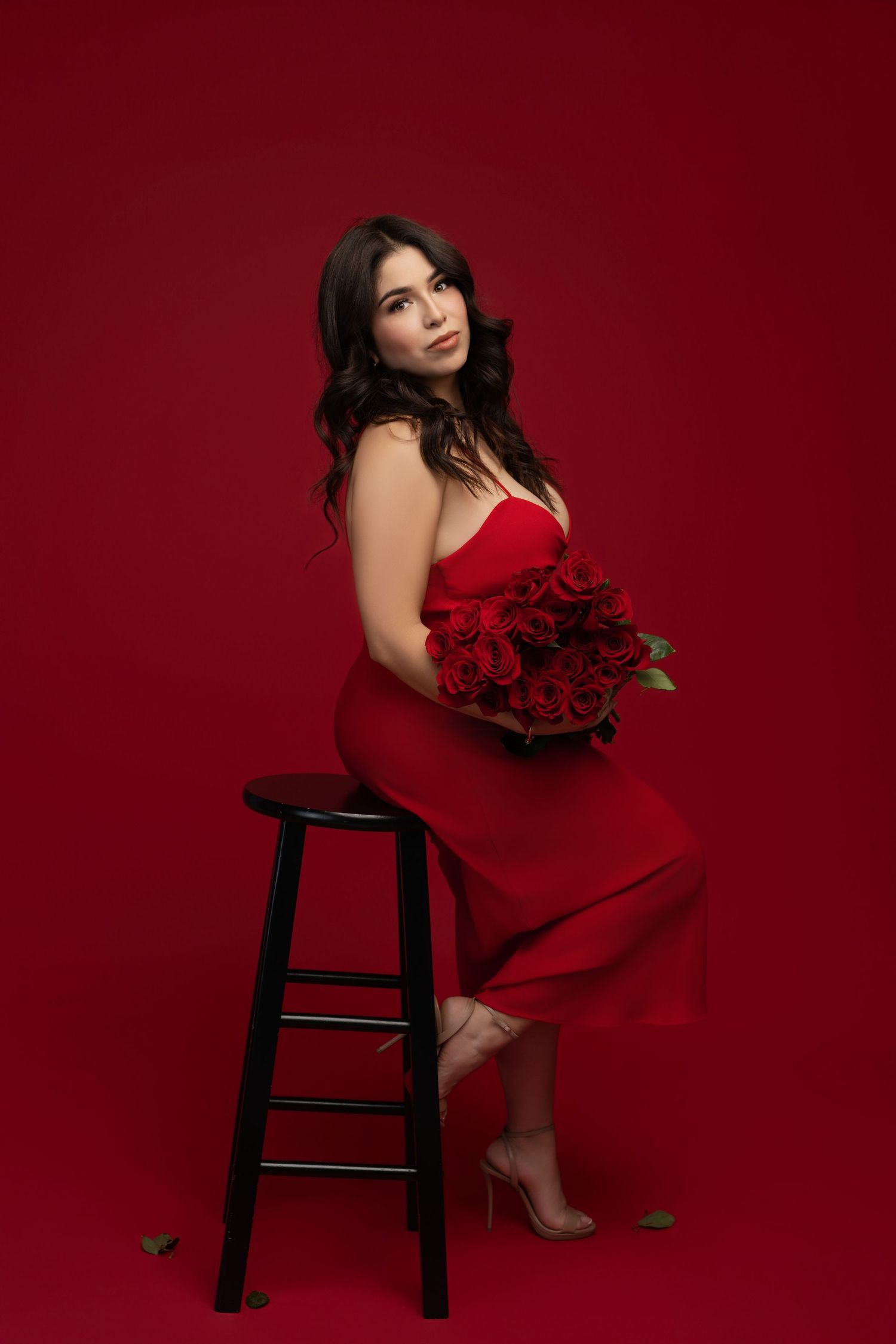 Woman in a red satin dress and strappy heels sits on a black stool holding a bouquet of red roses against a red studio backdrop.