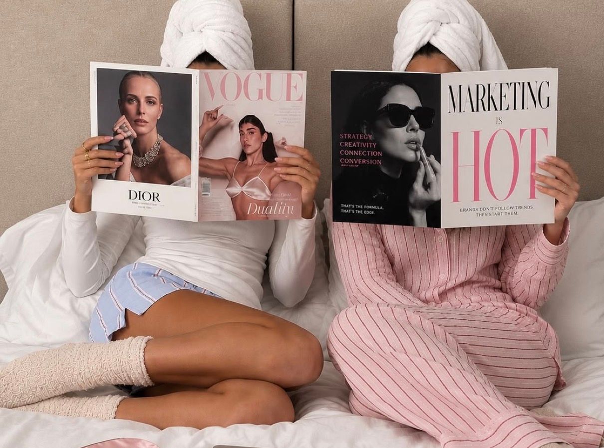 Two women in robes and towel wraps sit in bed holding Vogue and Marketing is Hot coffee table books.