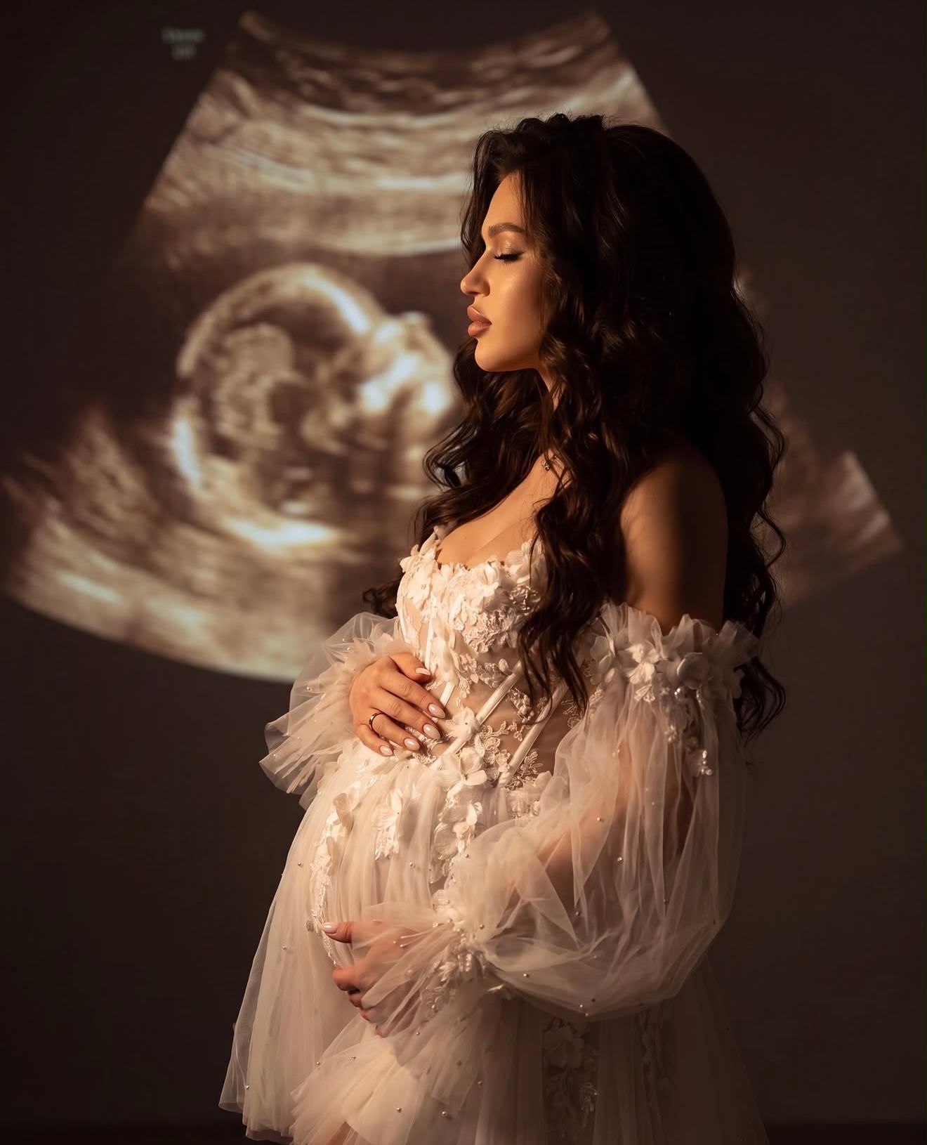 Pregnant woman in white floral tulle gown poses gracefully with ultrasound image projected behind her.