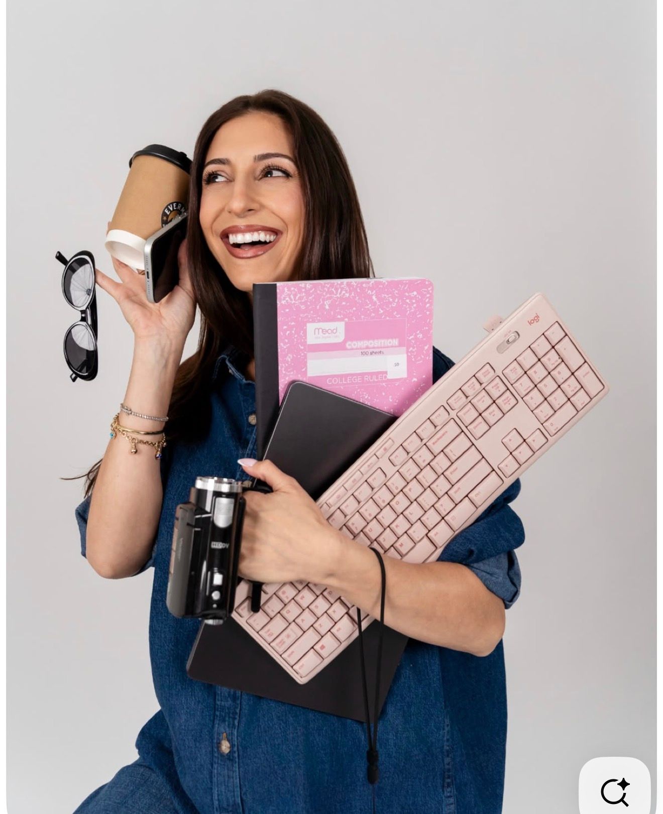 Smiling woman in denim shirt holding pink keyboard, notebook, laptop, coffee cup, and glasses against white background.
