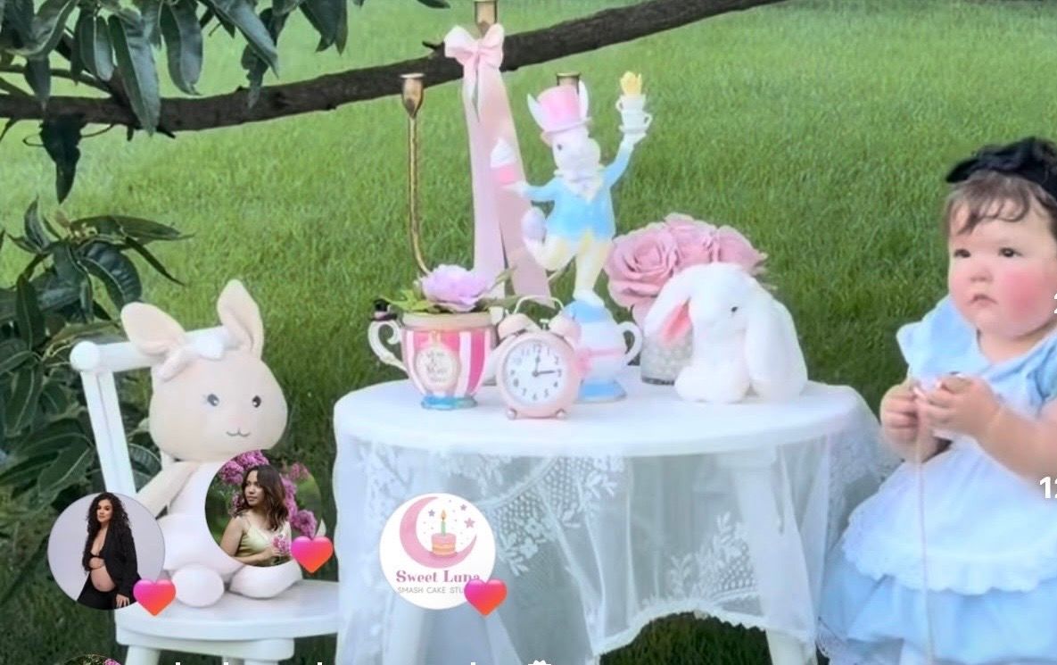 Alice in Wonderland themed first birthday party setup with white table, bunny toys, teacups, and pink roses on green lawn.