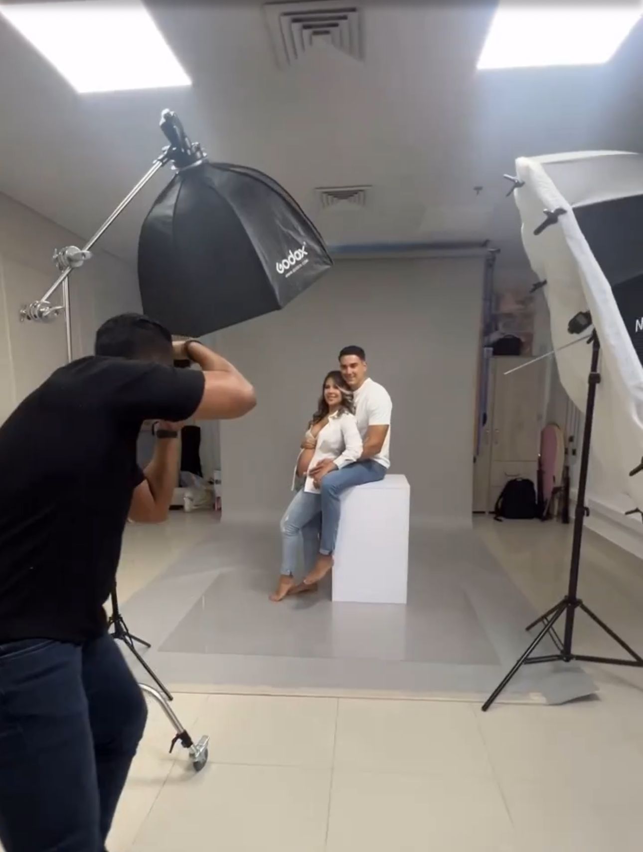 Photographer capturing couple posing on white cube in professional studio with Godox lighting.